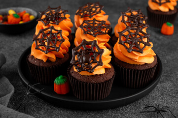 Halloween chocolate cupcakes with orange frosting and chocolate spider web on a dark background. Festive dessert. Funny food idea. Copy space.