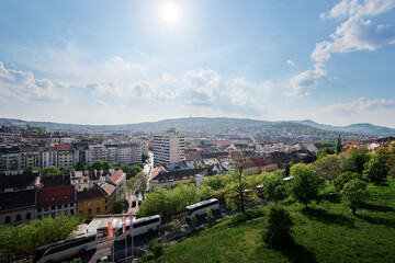 A scenic view of a cityscape with a mix of modern and historic buildings, surrounded by lush greenery and hills in the background under a partly cloudy sky.