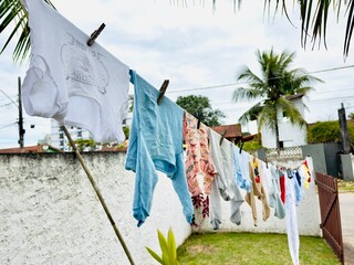 Roupas infantis coloridas a secar no varal num quintal tropical. Uma cena cotidiana, simples e familiar, com uma atmosfera de calor e vida doméstica.