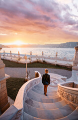 A woman in elegant attire walks down ornamental steps overlooking a serene waterfront during a...