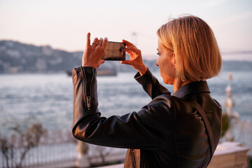 A woman captures a scenic view by the water at sunset, surrounded by serene landscapes. Woman...