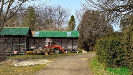 Autumn countryside road with red tractor and firewood near barn on sunny day, rural landscape with bare trees, green grass and warm seasonal light