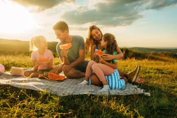 A young couple with their daughters eat watermelon at sunset while talking and enjoying the fresh...