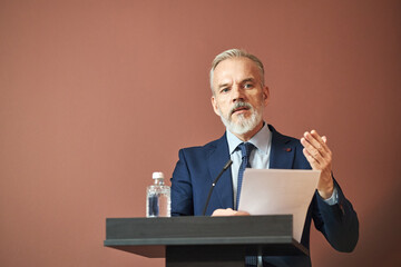 Middle aged Caucasian man standing at podium delivering speech, holding paper in one hand and gesturing with other, water bottle on podium, looking slightly past camera