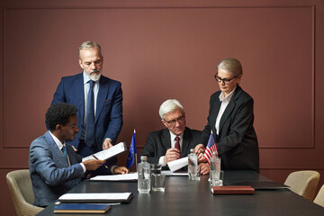 Group of middle aged and senior politicians including Black man, Caucasian man, Caucasian woman reviewing and signing documents at conference table with European and American flags visible