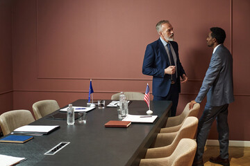 Middle aged Caucasian man and young adult Black man standing near conference table, discussing...