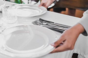 Close-up of elegant table setting with person placing a white plate on a white tablecloth, featuring glassware, cutlery, and napkins in a formal dining environment.