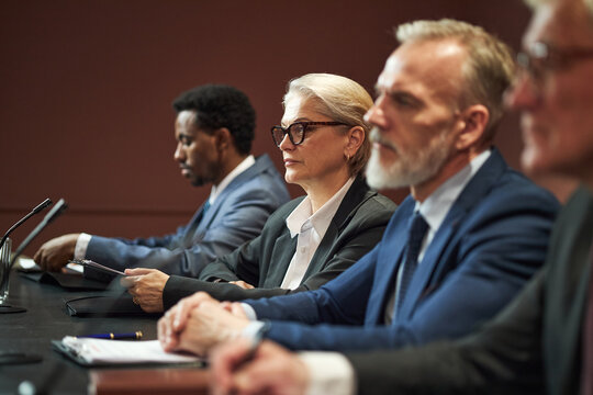 Middle aged Caucasian woman and middle aged Caucasian man sitting at conference table with Black man and another man, participating in political meeting, microphones visible - Powered by Adobe