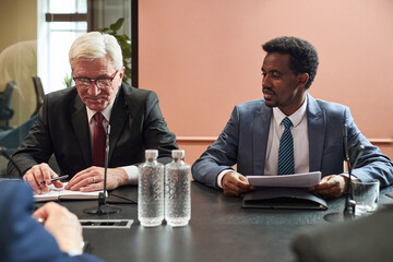 Caucasian senior man and Black middle aged man sitting at conference table discussing documents...