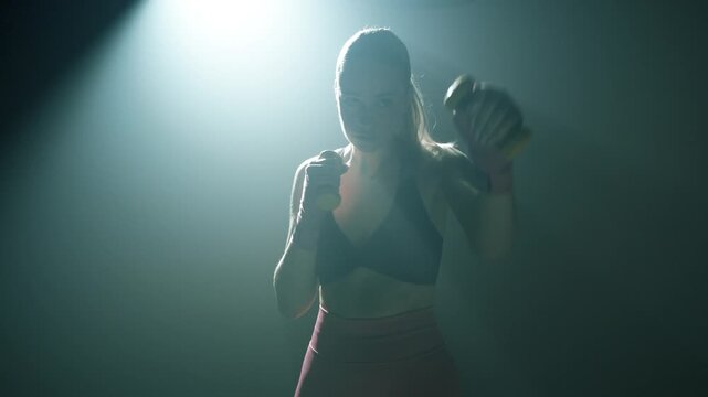 Zbliżenie na bokserkę shadow boxing z ż&oacute;łtymi ciężarkami w dłoniach, filmowe oświetlenie od tyłu, ciemna sala treningowa, koncentracja, siła i elegancja ruchu w slow motion