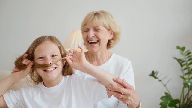  Funny and warm moment: the girl playfully makes a moustache out of her hair while the grandmother peeks over her shoulder, laughing. Captures family joy and spontaneous affection. High quality 4k