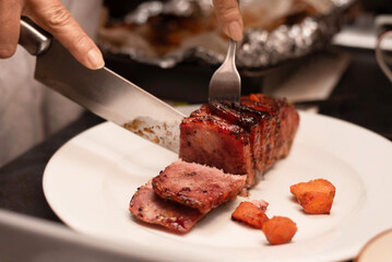 A sliced piece of grilled meat being served with fork and knife, accompanied by roasted vegetables. Woman cutting meat at home