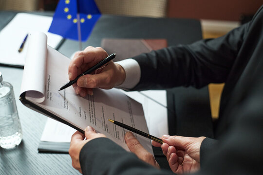 Caucasian middle aged men discussing official documents at table, one man holding clipboard while another pointing at paper, European Union flag visible in background, business meeting - Powered by Adobe