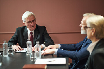 Senior Caucasian man in suit speaking during meeting with middle aged Caucasian man and middle aged...