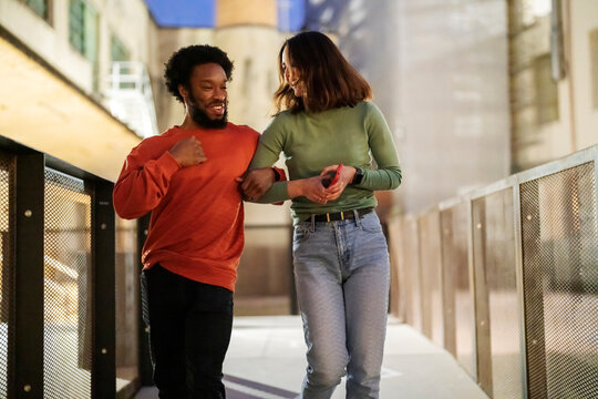 Happy black man in casual clothes smiling and telling funny story to girlfriend while walking on bridge on city street in evening.