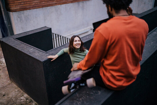 Happy woman with hands in pockets smiling and looking at black male skater while leaning on border outside brick building in evening on city street.