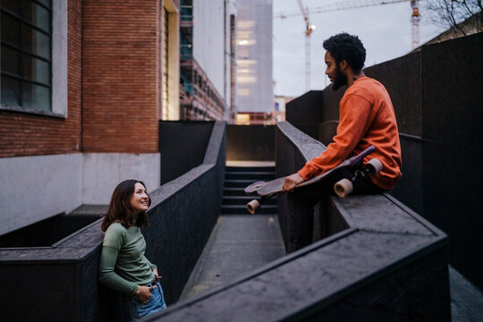 Happy woman with hands in pockets smiling and looking at black male skater while leaning on border outside brick building in evening on city street.