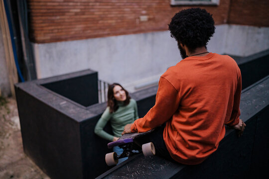 Happy woman with hands in pockets smiling and looking at black male skater while leaning on border outside brick building in evening on city street.