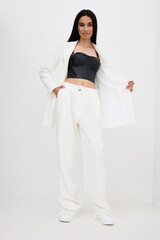 woman in white suit and black corset posing confidently with hands in pockets against plain background, stylish modern outfit, minimalistic fashion portrait