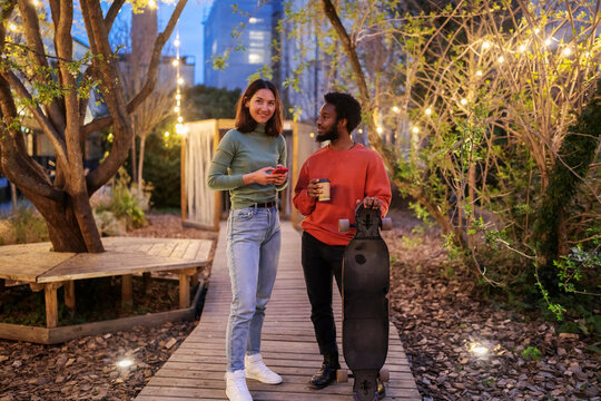 Happy woman with hands in pockets smiling and looking at black male skater while leaning on border outside brick building in evening on city street.