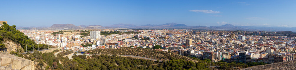 Panoramic view of Alicante, Spain, showcasing a vibrant cityscape with residential and commercial buildings, surrounded by hills and mountains under a clear blue sky.