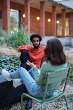 Happy African American male in casual clothes smiling and gesticulating while chilling in yard of apartment building and telling funny story to girlfriend in evening.