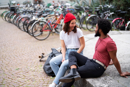 Happy diverse man and woman in casual clothes smiling and looking at each other while sitting on concrete border near bicycles on weekend day in city.