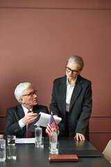 Caucasian senior man sitting at table discussing documents with Caucasian senior woman standing...