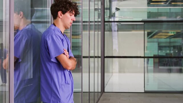 Exhausted young male doctor wearing scrubs sitting on floor in hospital corridor - shot in slow motion