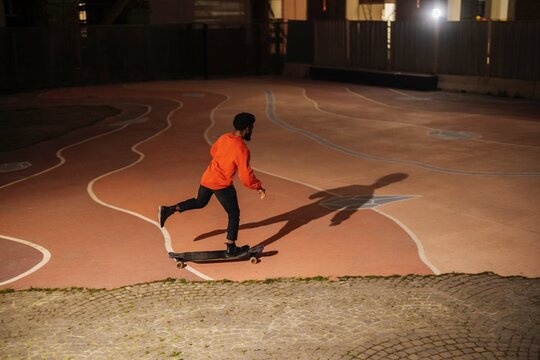 High angle of African American man in casual clothes riding longboard in dark skate park at night in city.