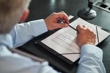 Senior Caucasian man sitting at desk reviewing official documents, holding pen in hands, preparing...