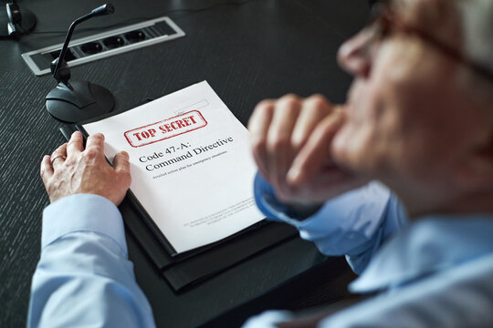 Senior Caucasian man sitting at conference table holding classified document labeled top secret, resting hand on folder, appearing thoughtful, microphone visible nearby