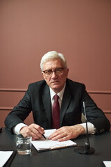 Portrait of senior Caucasian man wearing suit and glasses sitting at desk holding pen and paper,...