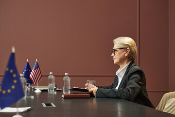 Middle aged Caucasian woman sitting at conference table holding glass, participating in diplomatic...