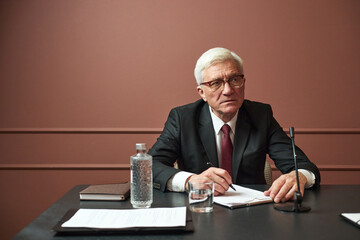 Senior Caucasian man wearing suit sitting at desk writing on notepad during meeting with documents,...