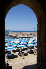 View of beach with blue umbrellas and sun loungers through stone archway at seaside harbor in sunny...