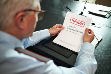 Senior Caucasian man reading classified document marked top secret at conference table, holding...