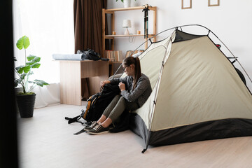 Female Backpacker in eyeglasses and casual clothes sitting into tent with backpack while preparing...