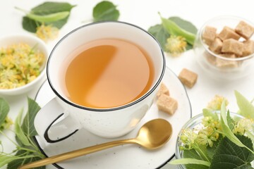 Tasty linden tea in cup, spoon, sugar cubes, leaves and flowers on white table, closeup