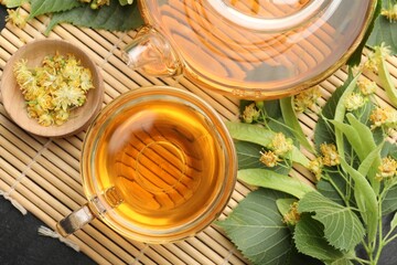 Tasty linden tea in cup, teapot, leaves and flowers on dark table, top view