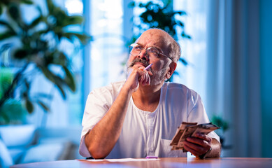 Elderly Indian man manages budget by counting cash and noting expenses at home