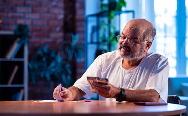 Elderly Indian man manages budget by counting cash and noting expenses at home