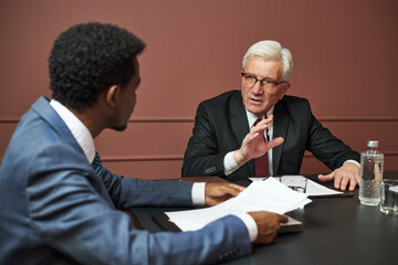 Middle aged Black man and senior Caucasian man discussing documents at table, senior man gesturing...