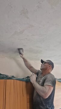 An elderly craftsman wearing glasses uses a sanding mesh tool to smooth plaster on a ceiling, working with one hand above his head. He is entirely covered in a thick layer of white dust from sanding