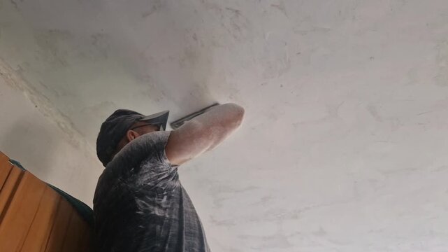 White sanding dust falls in dense veils from the ceiling as a skilled man works overhead with a manual sanding tool, smoothing the plastered surface during a room renovation