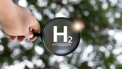 Hand holding magnifying glass zooming in on H2 hydrogen text with green nature background,...