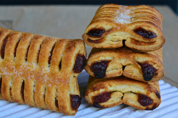 A stack of golden-brown, flaky pastries with visible dark filling, lightly dusted with sugar, stands beside another pastry on a white textured surface, all against a blurred background