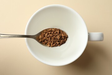 Spoon with instant coffee above cup on beige background, top view