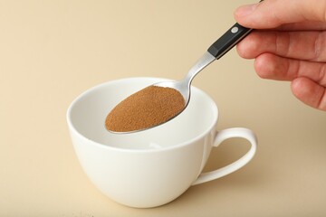 Woman putting instant coffee into cup on beige background, closeup