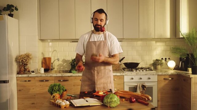 Young man in headphones juggling apples in the kitchen, wearing an apron and headphones. A playful moment during cooking at home. Relaxation, healthy lifestyle, fun cooking, mindfulness, routine